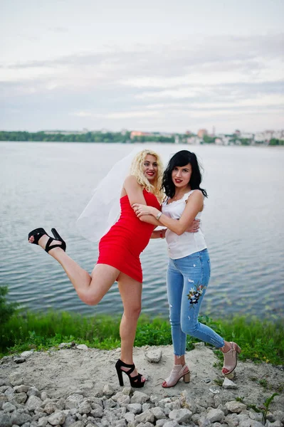 Impressionante noiva em vestido vermelho posando com sua dama de honra no lago — Fotografia de Stock
