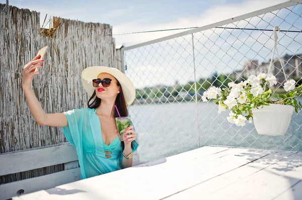 Sweet young lady wearing hat and sunglasses with a cocktail usin