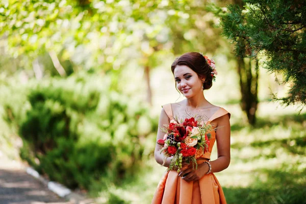 Привлекательная подружка невесты в оранжевом платье, позирующая с свадебным букетом — стоковое фото