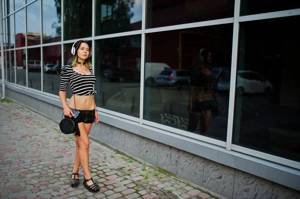 Chica llevar en pantalones cortos con auriculares grandes contra grandes ventanas bu —  Fotos de Stock