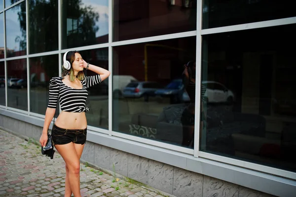 Chica llevar en pantalones cortos con auriculares grandes contra grandes ventanas bu — Foto de Stock