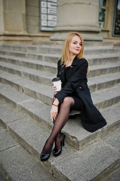 Elegant blond flicka bära på svart poserar på gatorna i staden, sitta — Stockfoto