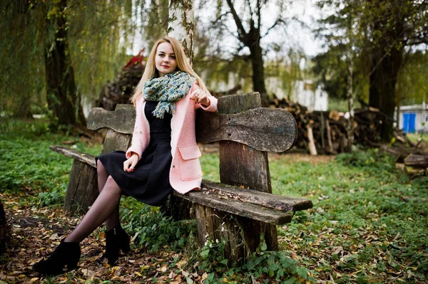 Mladá blondýnka v růžový kabát sedí na lavičce položené proti wo — Stock fotografie