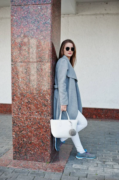 Mädchen im grauen Mantel mit Sonnenbrille und Handtasche bleibt gegen Wand. — Stockfoto