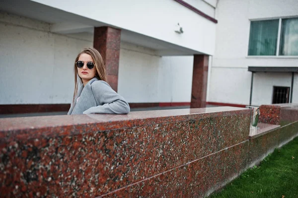 Mädchen im grauen Mantel mit Sonnenbrille und Handtasche bleibt gegen Wand. — Stockfoto