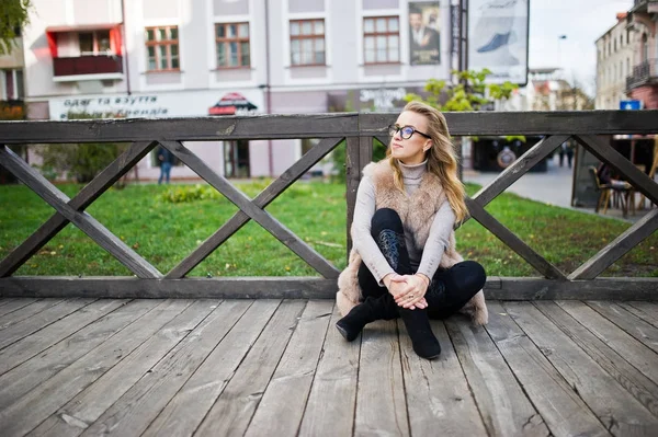 Blondes Mädchen im Pelzmantel sitzt auf Holzboden im Freien. — Stockfoto