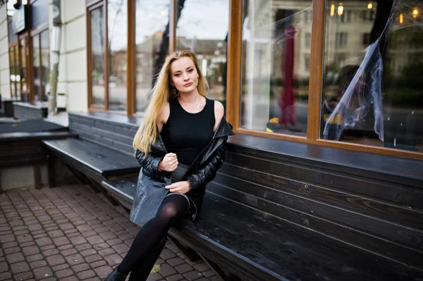 Blonde fashionable girl in long black leather coat sitting on be — Stock Photo, Image