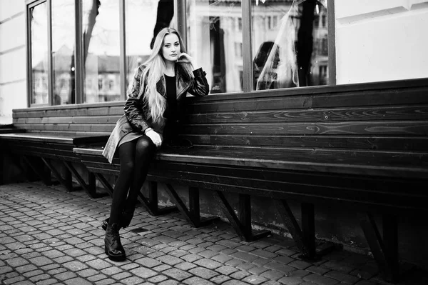 Blonde fashionable girl in long black leather coat sitting on be — Stock Photo, Image