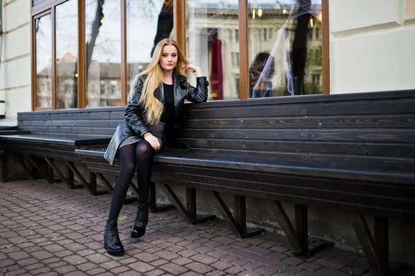 Blond fashionabla flicka i lång svart läder kappa sitter på vara — Stockfoto