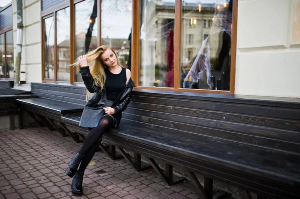 Blonde fashionable girl in long black leather coat sitting on be — Stock Photo, Image