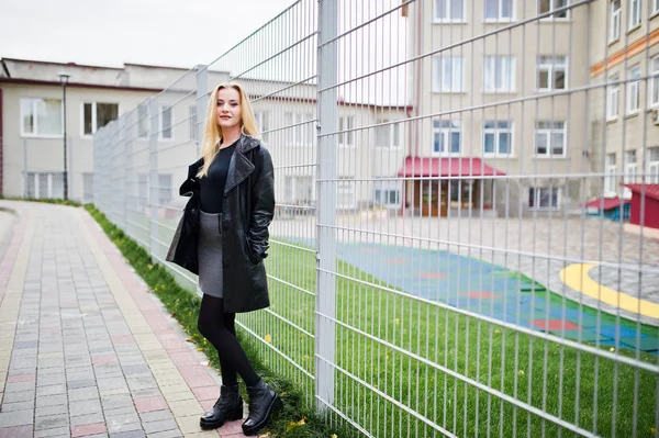 Blonde fashionable girl in long black leather coat against iron — Stock Photo, Image