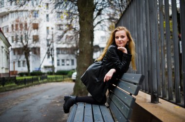 Uzun siyah deri montlu sarışın moda kız benc üzerinde poz verdi.