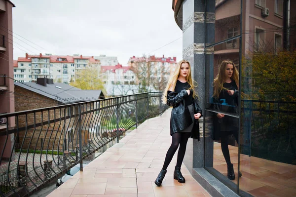 Blondes modisches Mädchen im langen schwarzen Ledermantel posierte gegen — Stockfoto