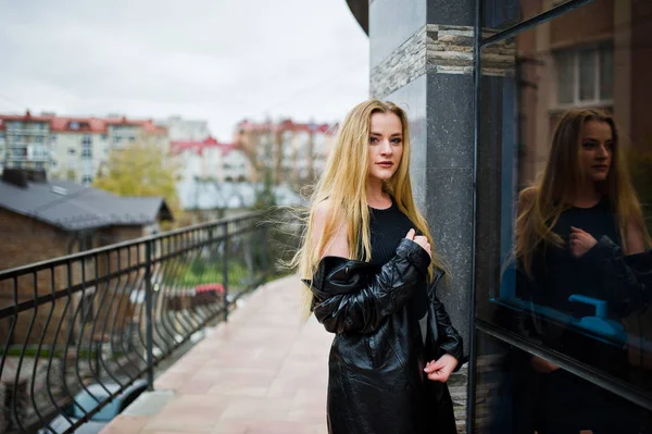 Blondes modisches Mädchen im langen schwarzen Ledermantel posierte gegen — Stockfoto