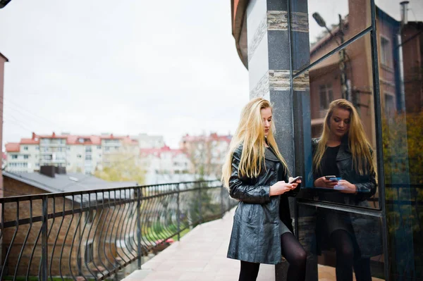 Blondes modisches Mädchen im langen schwarzen Ledermantel posierte gegen — Stockfoto