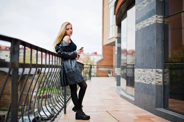 Blonde fashionable girl in long black leather coat posed against — Stock Photo, Image