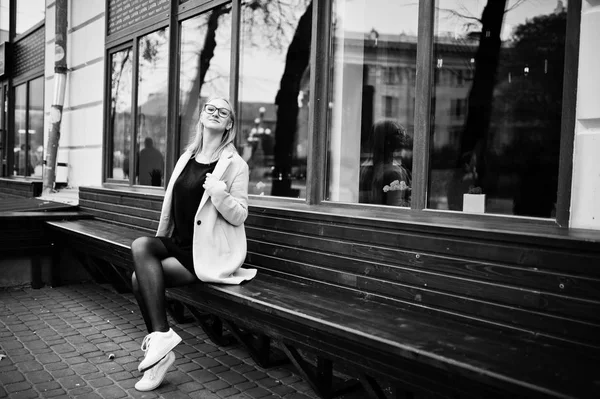 Blonde girl at glasses and pink coat, black tunic sitting on ben — Stock Photo, Image