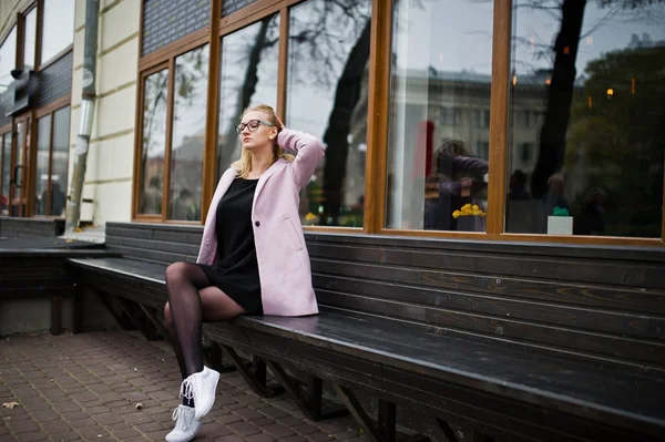 Blondes Mädchen mit Brille und rosa Mantel, schwarze Tunika sitzt auf Ben — Stockfoto