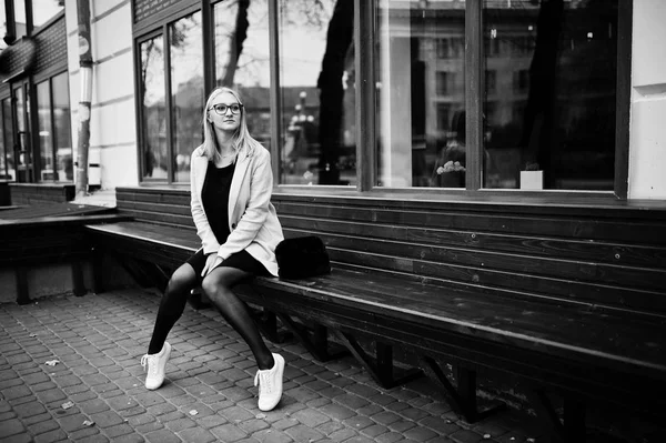 Blonde fille à lunettes et manteau rose, tunique noire assise sur ben — Photo