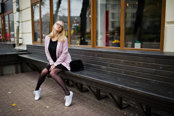 Blond flicka på glasögon och rosa kappa, svart tunika sitter på ben — Stockfoto