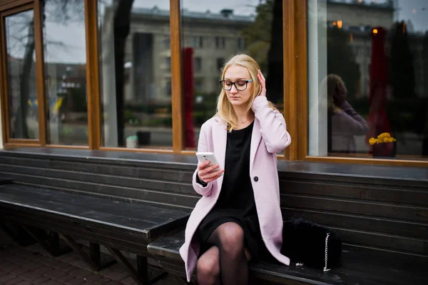 Blonde girl at glasses and pink coat, black tunic sitting on ben — Zdjęcie stockowe