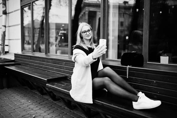 Blonde girl at glasses and pink coat, black tunic sitting on ben — Stock Photo, Image