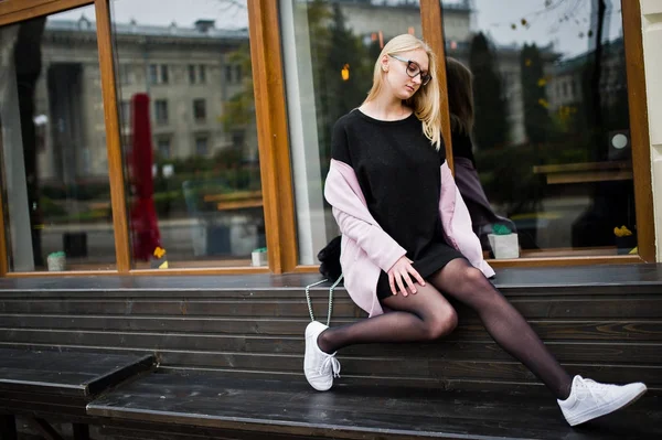 Blonde girl at glasses and pink coat, black tunic sitting on ben — Zdjęcie stockowe