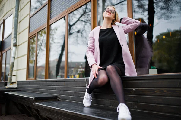 Blonde girl at glasses and pink coat, black tunic sitting on ben — Stock Photo, Image