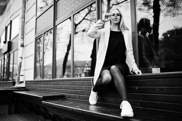 Blonde girl at glasses and pink coat, black tunic sitting on ben — Stock Photo, Image