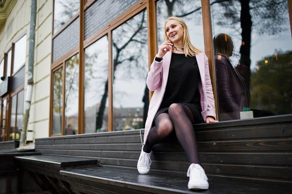 Blonde girl at glasses and pink coat, black tunic sitting on ben — Stock Photo, Image