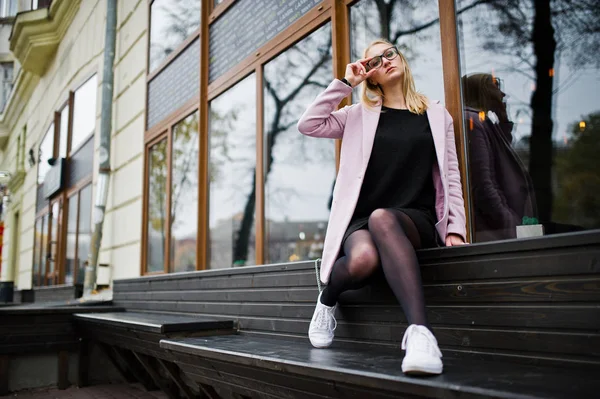 Blonde girl at glasses and pink coat, black tunic sitting on ben — Stock Photo, Image