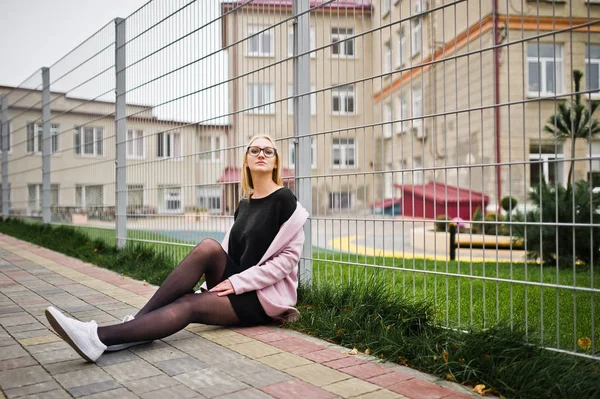 Blond flicka på glasögon och rosa kappa, svart tunika sittande agains — Stockfoto