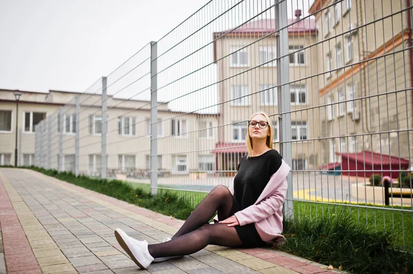 Blonde girl at glasses and pink coat, black tunic sitting agains — Stock Photo, Image