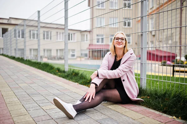 Blond flicka på glasögon och rosa kappa, svart tunika sittande agains — Stockfoto