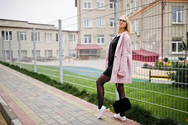 Blondes Mädchen mit Brille und rosa Mantel, schwarzer Tunika und Handtasche — Stockfoto