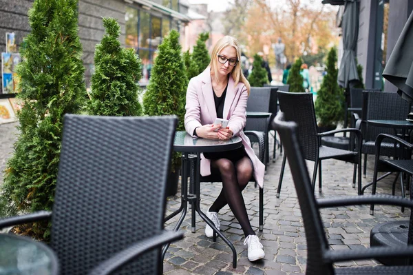 Ξανθό κορίτσι σε γυαλιά και ροζ παλτό, μαύρο χιτώνα που κάθεται σε καρτέλα — Φωτογραφία Αρχείου