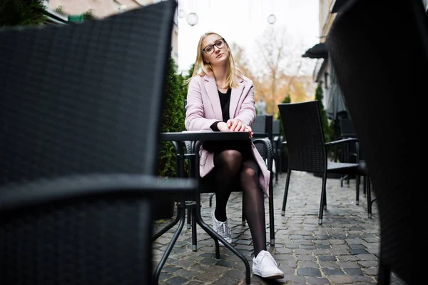 Blond flicka på glasögon och rosa kappa, svart tunika sitter på fliken — Stockfoto