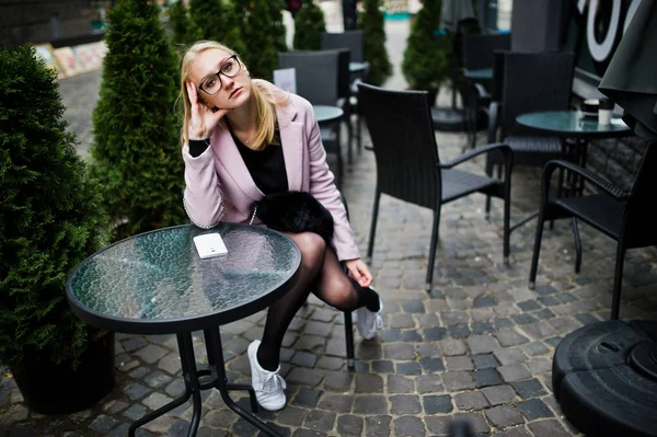 Blonde girl at glasses and pink coat, black tunic sitting at tab — Stock Photo, Image