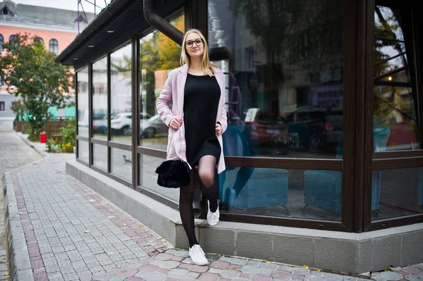 Ragazza bionda a occhiali e cappotto rosa, tunica nera contro finestra — Foto Stock
