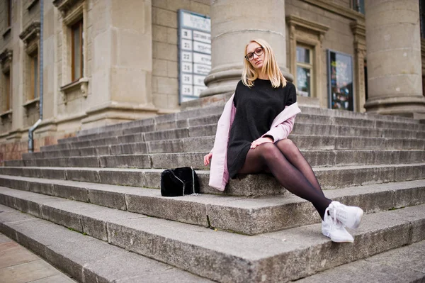 Blond flicka på glasögon och rosa kappa med handväska sitter på sta — Stockfoto