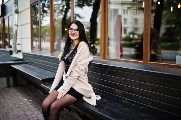 Schwarze Haare sexy Frau mit Brille und Mantel sitzt auf Bank. — Stockfoto