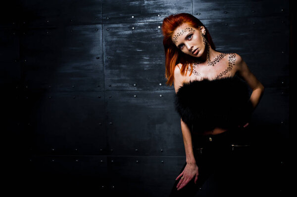 Fashion model red haired girl with originally make up like leopard predator against steel wall. Studio portrait.