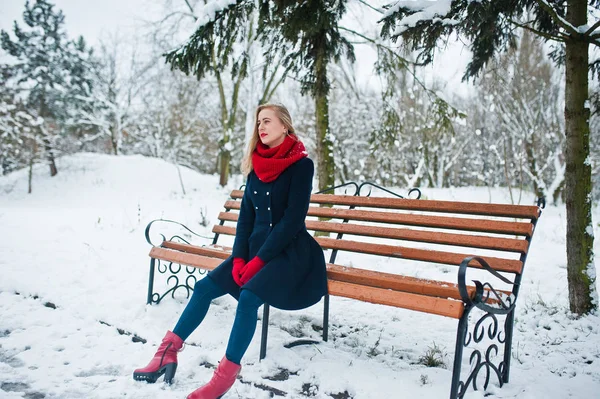 Блондинка в красном шарфе и пальто сидит на скамейке в зимний день — стоковое фото