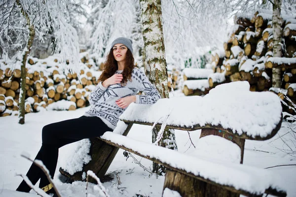 Menina encaracolado bonito em suéter e headwear sentado no banco na neve — Fotografia de Stock