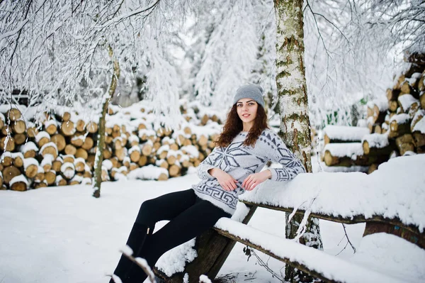 Χαριτωμένο σγουρά κορίτσι σε πουλόβερ και πιλοποιία, κάθεται στο παγκάκι στο χιόνι — Φωτογραφία Αρχείου