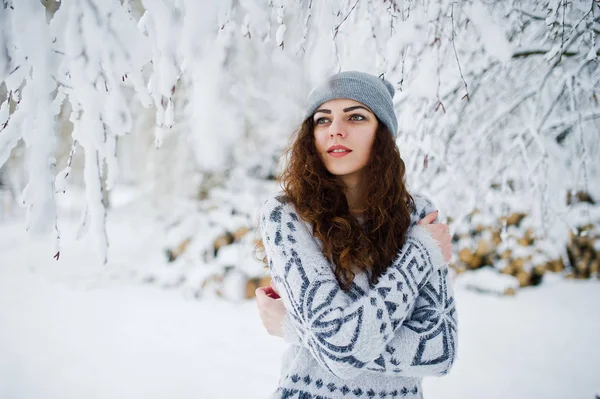 Carino ragazza riccia in maglione e copricapo al parco forestale innevato a — Foto Stock