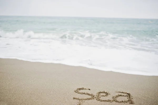 Zbliżenie zdjęcie piasku napis "morze" przez pieniaste fale. — Zdjęcie stockowe