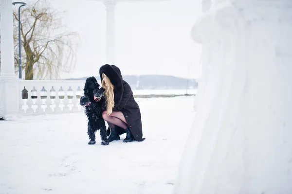 Menina loira elegância no casaco de pele abraços pequeno cão preto no inverno — Fotografia de Stock