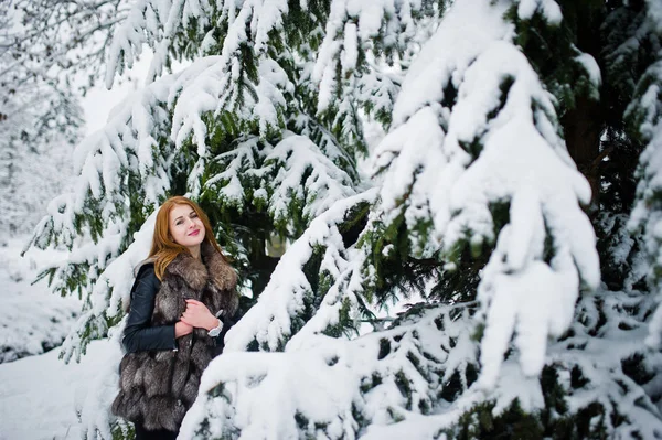 Menina de cabelos vermelhos em casaco de pele andando no parque nevado de inverno . — Fotografia de Stock