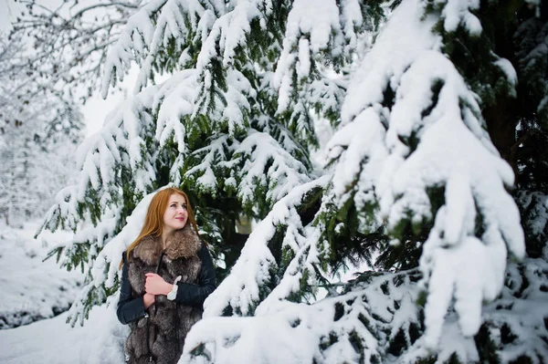 Menina de cabelos vermelhos em casaco de pele andando no parque nevado de inverno . — Fotografia de Stock